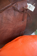 Distressed dark whiskey leather pouf with tie closure, beside orange leather with fan pattern.