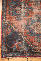 Antique Mehrevan rug corner with distressed burgundy field, geometric blue palmettes, and fringe.