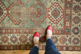 Feet on a distressed Antique Kazak rug with light green field and geometric patterns.