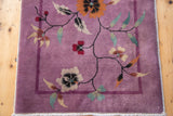 Mauve purple Art Deco rug corner with sprawling floral vines, thin banded border, and white fringe.
