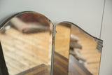 Upper right hinge and curved dark wood edge of an antique tri-folding mirror with aged glass.