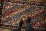 Antique Caucasian rug, geometric floral motifs in cinnabar red, blue, and cream beneath black boots.