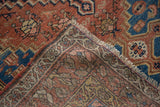 Corner of antique distressed Malayer rug, showcasing worn brick red, blue geometric patterns and fringe.