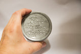 Hand holding the aged zinc lid of a vintage Ball Jelly Glass jar, with embossed text.