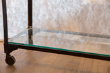 Antique industrial table's glass bottom shelf, dark cast iron, and caster wheel.