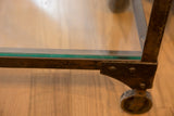 Antique industrial table's aged cast iron frame, bolted corner, glass shelf edge, and wheel.