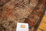 Distressed texture of antique Malayer rug, showing worn madder red, blue Herati pattern, and tattered edge.