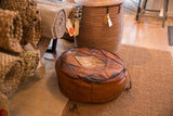 Vintage leather pouf with patchwork top, lighter central panel, and aged brown patina.