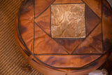 Side of vintage leather pouf with dark piping, varied brown patina, and original tassel.