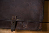 Distressed dark brown leather messenger bag flap corner with light stitching and wear.