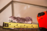 Purple amethyst crystal cluster, its rough texture measured by a yellow tape.