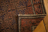 Distressed Belouch rug's folded corner, showing secured fringe and geometric gul pattern in warm brown, red, and navy.