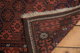 Folded corner of the vintage Afghan rug, showing geometric gul motif, crab border, goat hair selvage, and ivory fringe in brick red and charcoal.