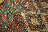 Geometric diamond and boteh patterns in madder red, navy, and ivory on vintage Caucasian rug, showing wear.