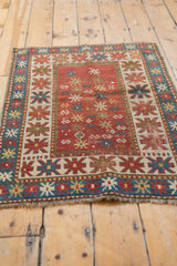 Antique Kazak mat with terra cotta red field, blue starburst borders, and playful motifs.