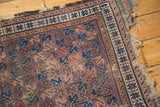 Corner of antique Belouch rug with aubergine field, geometric plant patterns, and tattered fringe.