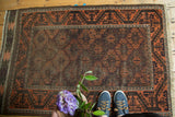 Corner of antique Belouch rug showing geometric lattice trellis and scrolling border in time-softened rust and brown.