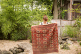 Antique Turkmen square rug held up, displaying dark red geometric gul motifs with worn fringe.