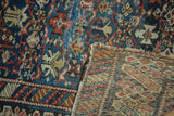 Antique Caucasian rug's folded corner, revealing worn navy blue and red geometric pile, visible foundation, and tattered edge.