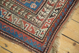 Vintage Caucasian rug corner with geometric red, blue, and beige borders, showing a tattered edge.