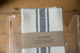Handwoven napkin showing cream and black stripes, woven cotton texture, and brand label.