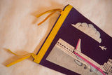 Upper corner of vintage airport felt pennant, yellow binding, purple background, white cloud, and plane.
