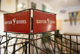 Vintage Bantam Books red metal sign with white rooster logo on display rack.