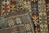 Folded antique Caucasian rug corner reveals geometric starbursts, tattered fringe, and hand knotted back.