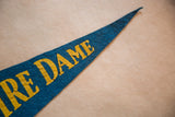Vintage blue felt Notre Dame pennant, yellow 'DAME' lettering, visible hole near 'A'.