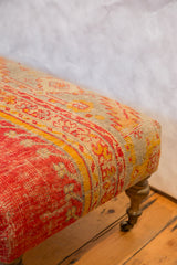 Ottoman upholstered in antique Turkish rug fragment with pink, yellow, aqua patterns, brass wheel.