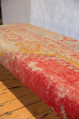 Side view of extra long ottoman upholstered with distressed antique Turkish rug fragment, faded pink, yellow.