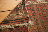 Antique Belouch rug corner with geometric ivory border, madder red field, and tattered fringe.