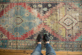 Feet on a vintage distressed Oushak runner, showing geometric patterns and worn texture in blue, red, and saffron yellow.