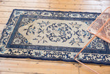 Ivory field of antique Peking rug, dark blue curvilinear design, running-T border, and visible edge wear.