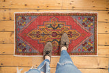 Distressed Oushak rug corner featuring worn brick red, dusty gold, and slate blue geometric patterns.