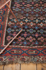 Antique Kurdish rug's worn charcoal geometric field, madder red border, and tattered fringe.