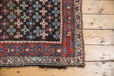 Corner of antique Kurdish rug, with geometric charcoal field, madder red borders, and worn tattered edge.
