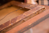 Antique painting frame's back corner, revealing aged reddish-brown wood and hanging wire.