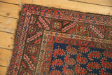 Corner detail of an antique Kurdish rug, showing geometric borders, brick red botehs, and tattered fringe.