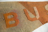 Detail of antique B U S felt pennant, orange and off-white letters, time-softened wool texture.