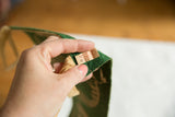 Hand holding vintage green felt pennant, showing aged paper tag and beige backing.