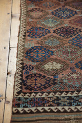 Corner of antique Belouch rug with geometric gul motifs, charcoal brown borders, and woven kilim end.