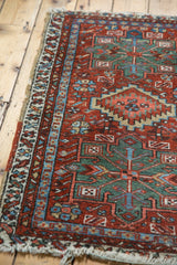 Corner of a vintage Karaja rug, highlighting brick red and mallard green geometric patterns with tattered fringe and intact selvage.