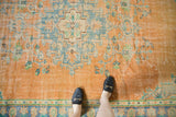 Feet on distressed Oushak rug, revealing faded tangerine, royal blue, mint floral motifs.