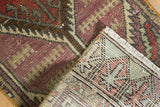 Distressed Oushak rug corner detail with geometric aubergine, rust, green patterns and secured fringe.