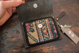 Open black leather wallet, displaying a geometric rug fragment in blue, red, beige with metal snap.