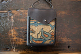 The front of a leather shoulder bag showing a woven rug fragment panel and snap closure.