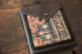 Close-up: dark pebbled leather flap with metal snap over a patterned rug fragment.