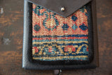 Hand-knotted rug fragment with geometric peach, muted blue, and red pattern, dark leather snap flap.