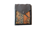 Dark pebbled leather flap, silver metal snap, and earthy geometric rug fragment.